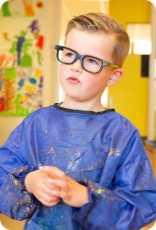 Jongen met bril en verf op zijn blauwe schilderschort kijkt nadenkend naar rechts.