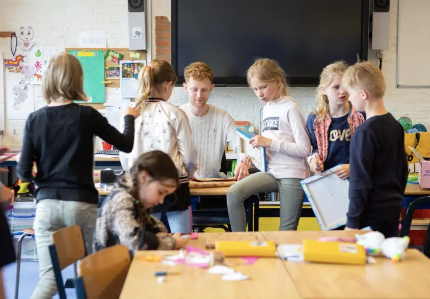 Kinderen in een klaslokaal bezig met creatieve activiteiten, begeleid door een leraar met rood haar.