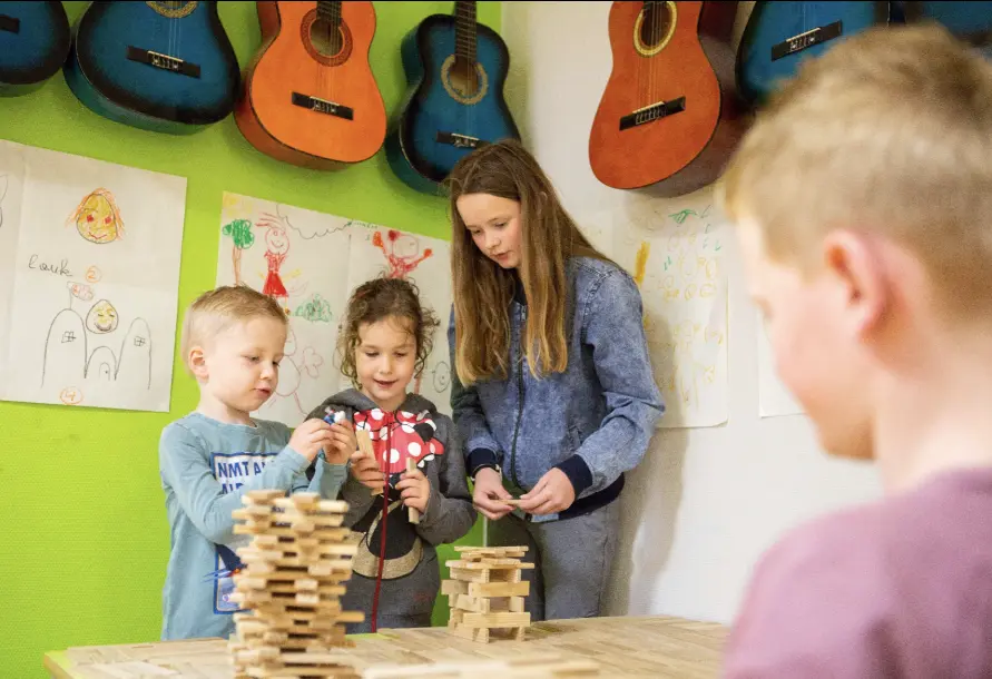 Drie kinderen bouwen met houten blokken aan een tafel, met gitaren en kindertekeningen aan de muur.