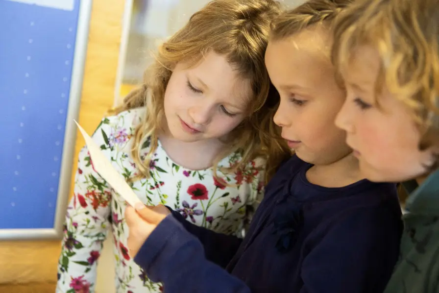 Drie kinderen lezen samen aandachtig een stuk papier in een binnenruimte.
