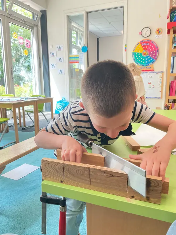 Jongen in een gestreept shirt zaagt een stuk hout op een groene tafel in een klaslokaal.