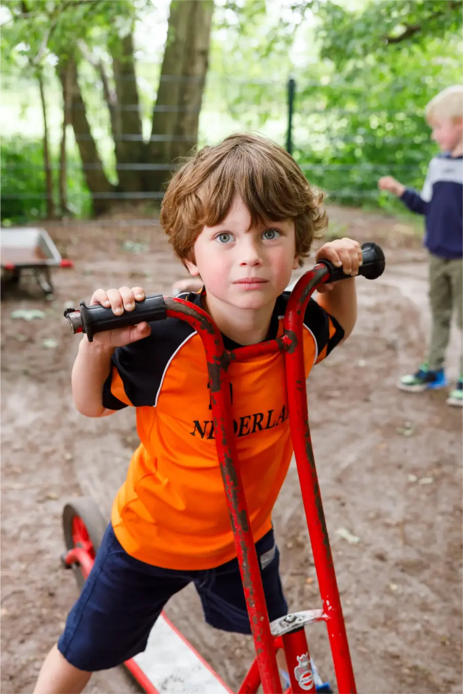 Jongen in oranje shirt met 'Nederland' vasthoudend aan het stuur van een rode step buiten.