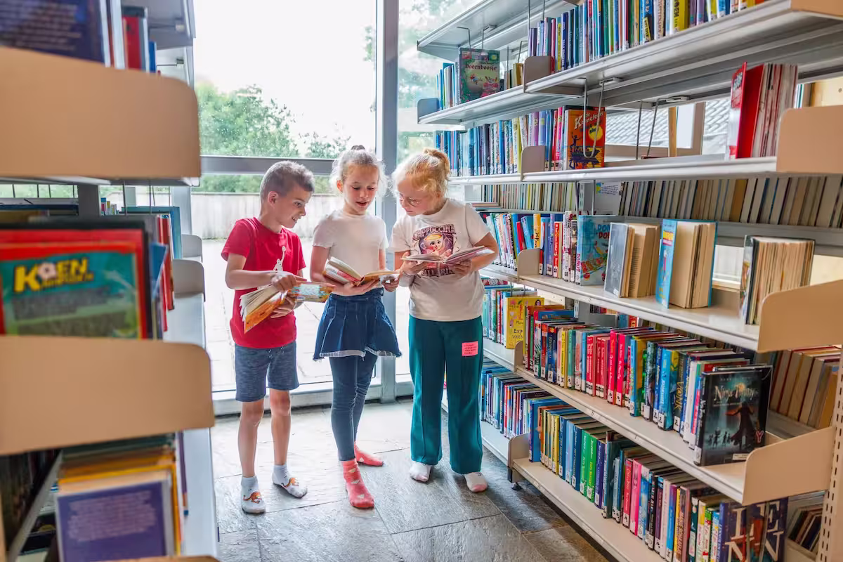 Drie kinderen lezen boeken samen tussen boekenplanken in een lichte bibliotheek.