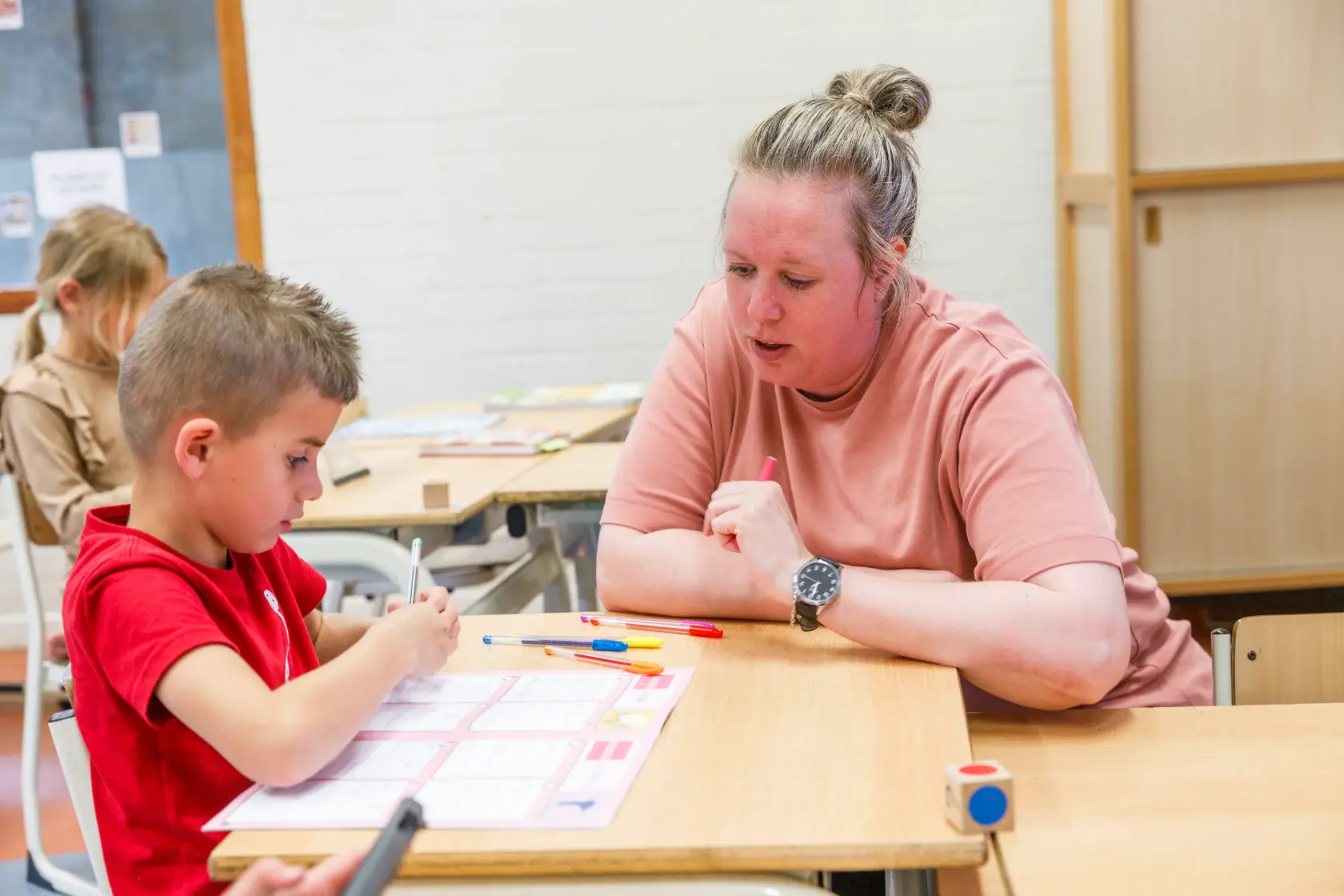 Leraar helpt jonge jongen in rood t-shirt met schoolwerk aan tafel in klaslokaal.