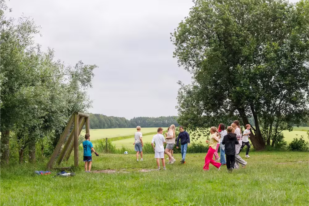 Kinderen spelen buiten voetbal in een groene veldomgeving met bomen en een houten doel.