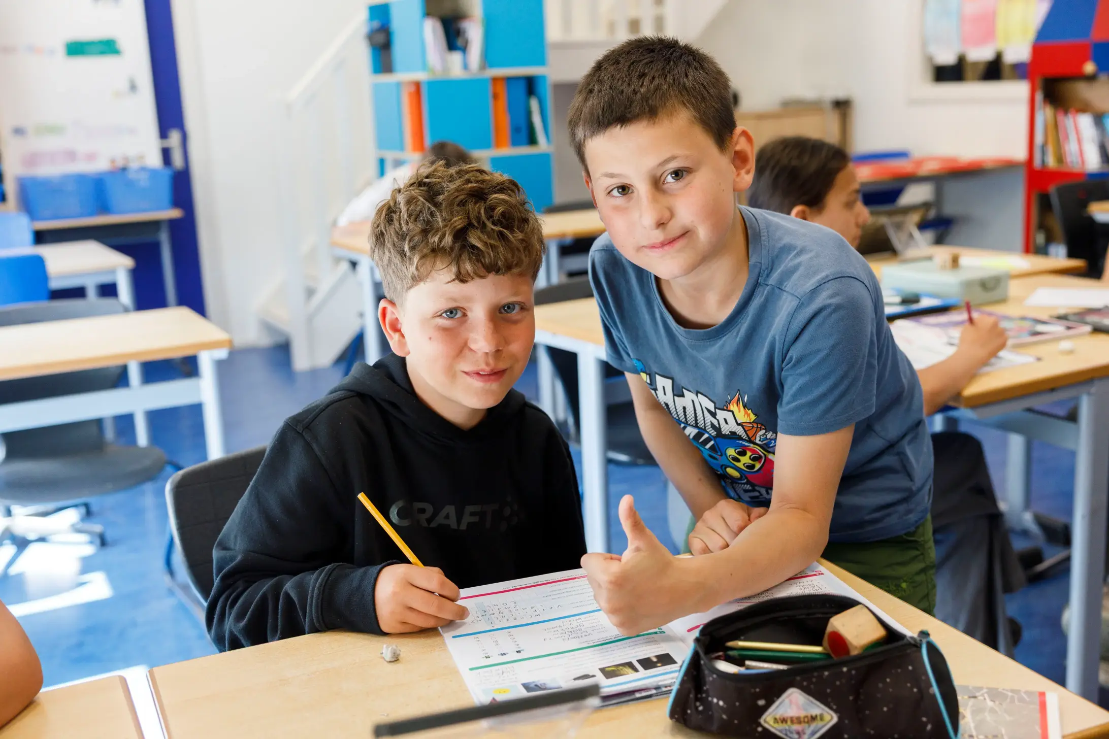 Twee jongens in een klaslokaal, één schrijft, de ander geeft duim omhoog bij het bureau.