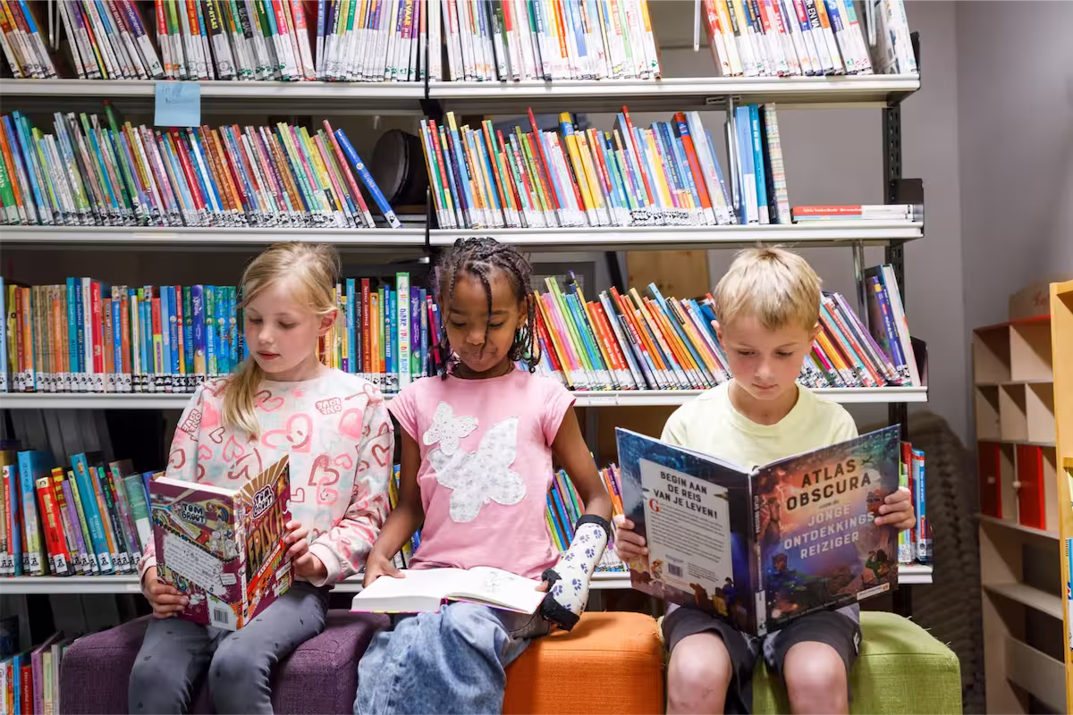Drie kinderen zitten te lezen in een bibliotheek met boekenplanken vol kleurrijke boeken achter hen.