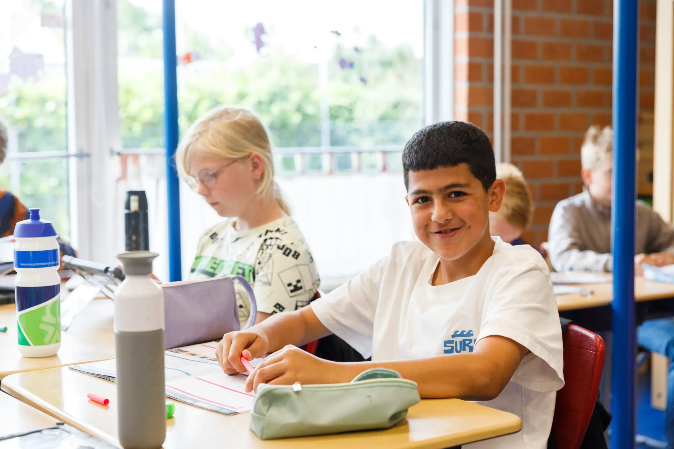 Jongen met wit shirt kijkt lachend naar camera terwijl hij in klas aan tafel werkt.