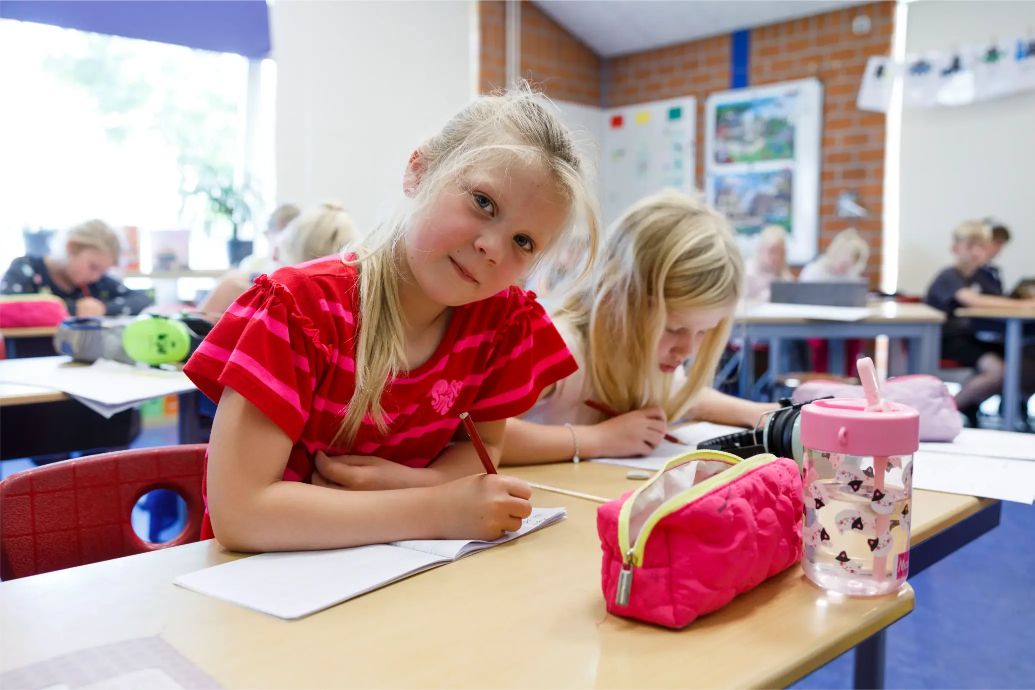 Twee meisjes schrijven zittend aan een tafel in een klaslokaal with pennen, etui en drinkbeker.