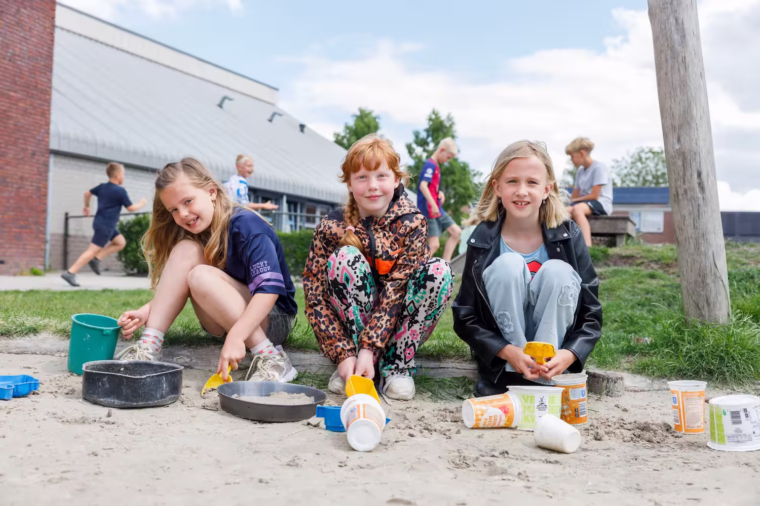 Drie meisjes spelen in het zand met emmers en scheppen op een zonnige dag buiten school.