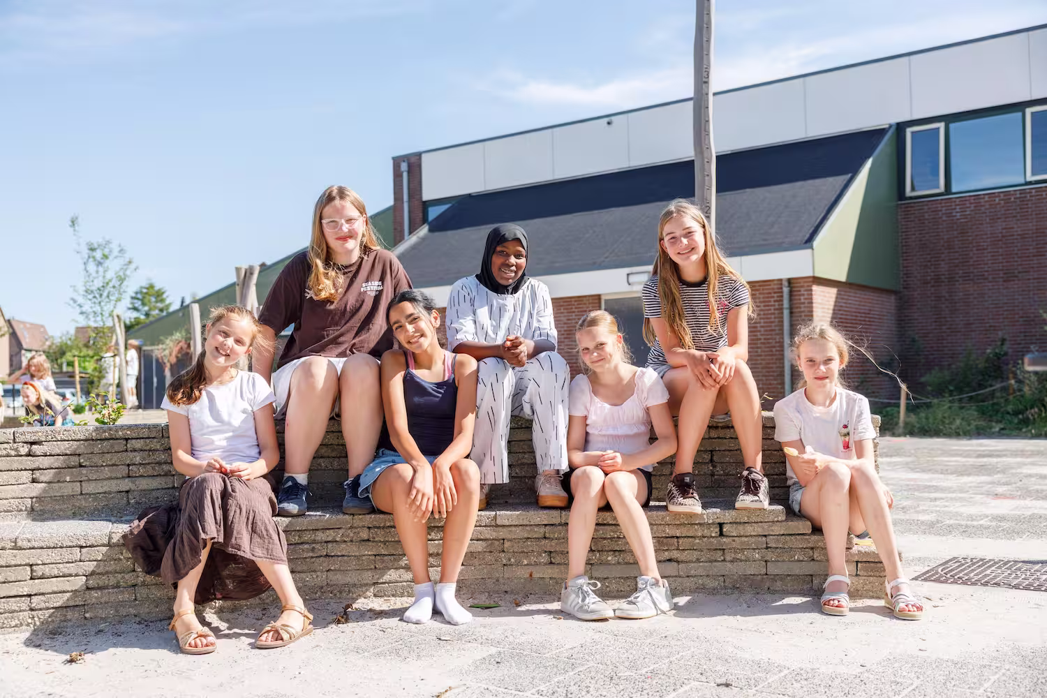 Zes meisjes zitten samen buiten in de zon op een lage stenen rand, lachend en ontspannen.