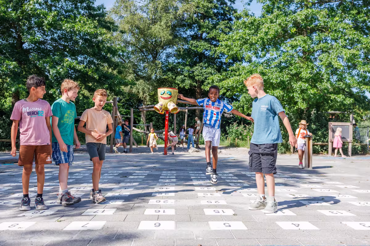 Vijf kinderen spelen op een speelplaats met een grote getallenvloer onder bomen op een zonnige dag.