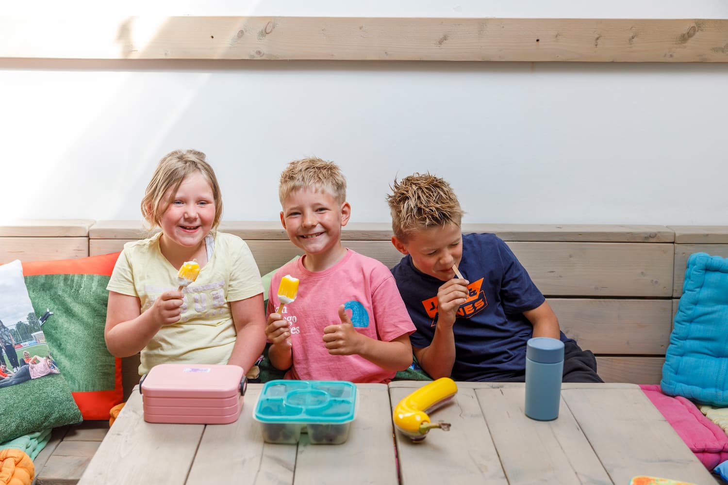 Drie kinderen zitten op een bank, twee eten ijsjes en een drinkt uit een beker aan een houten tafel.