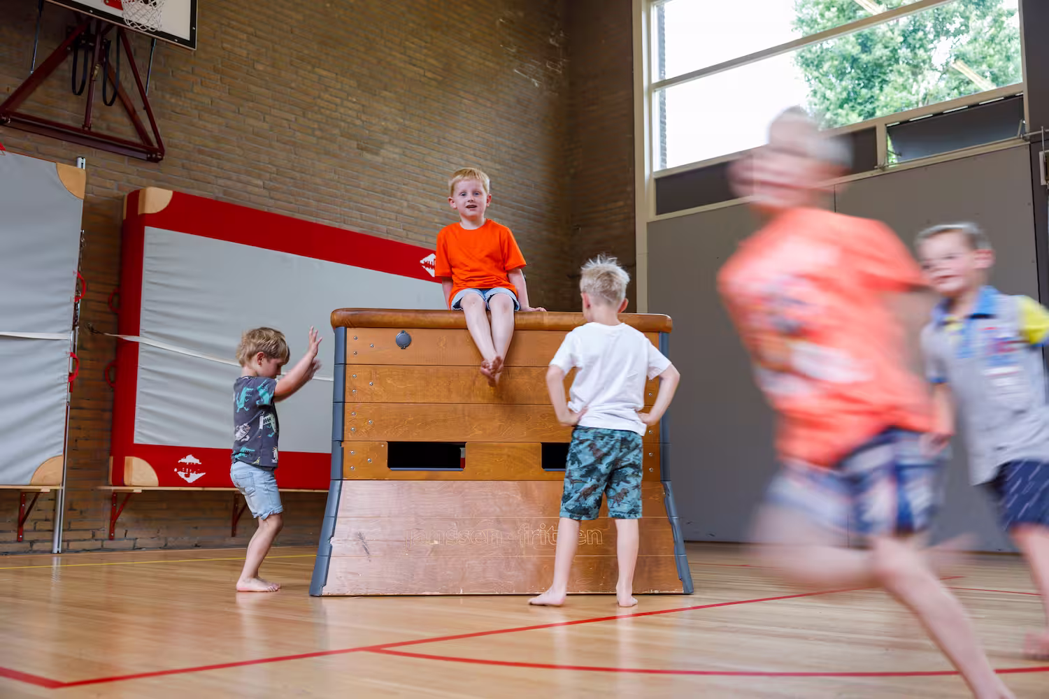 Kinderen spelen in een gymzaal met één kind zittend op een houten gymnastiekbok en anderen eromheen.