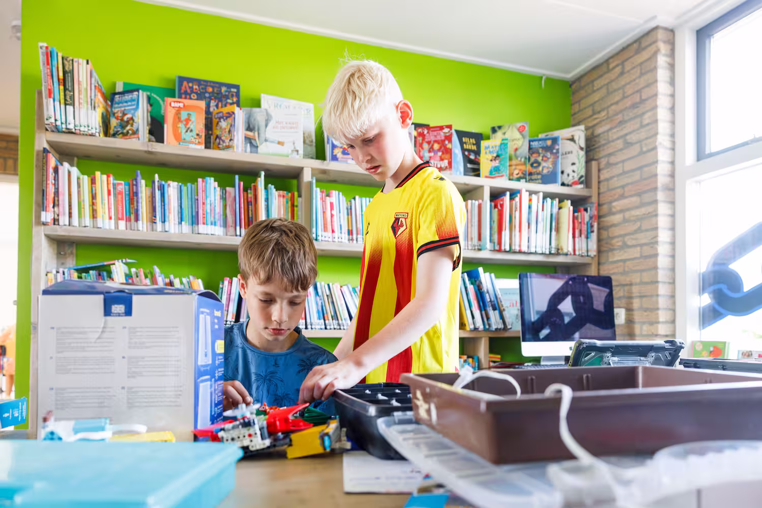 Twee jongens bouwen samen met LEGO in een kleurrijke bibliotheek vol boeken.