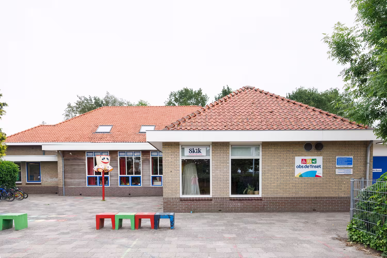Schoolgebouw met rood pannendak, kleurrijke banken en een speelplaats met een rode paal en figuur.