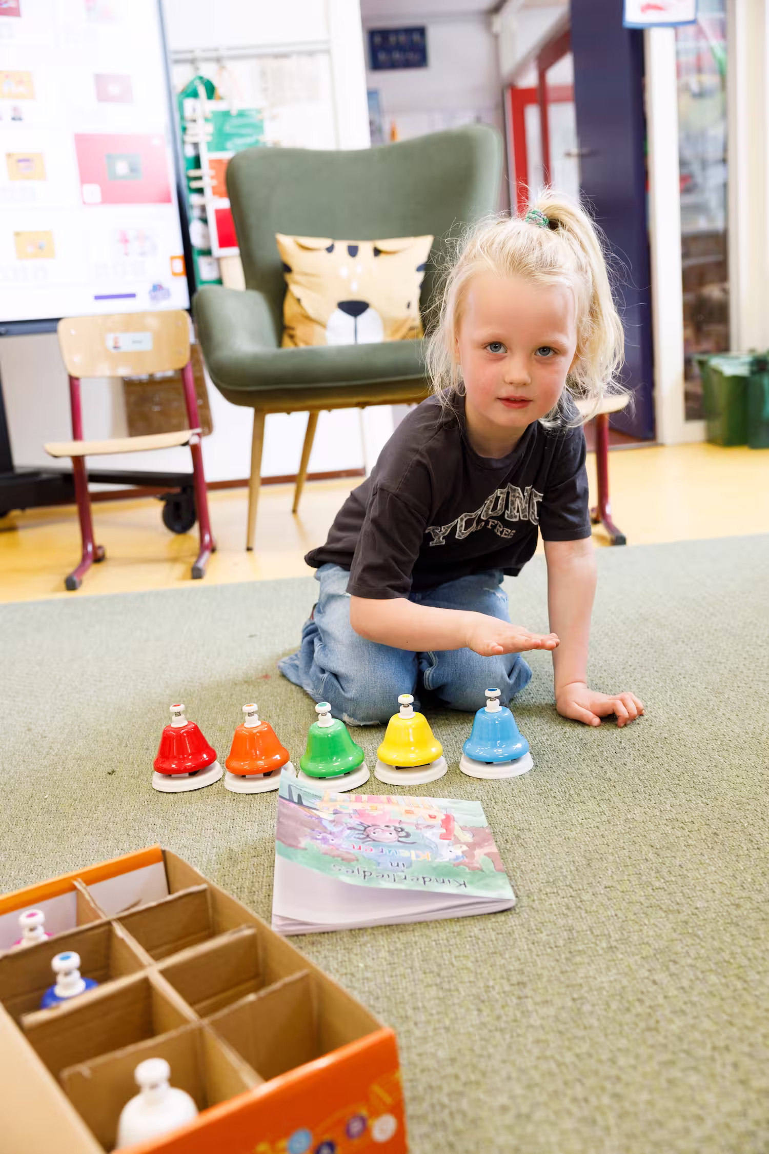 Kind speelt op de grond met gekleurde belletjes, met een boek en stoelen op de achtergrond.
