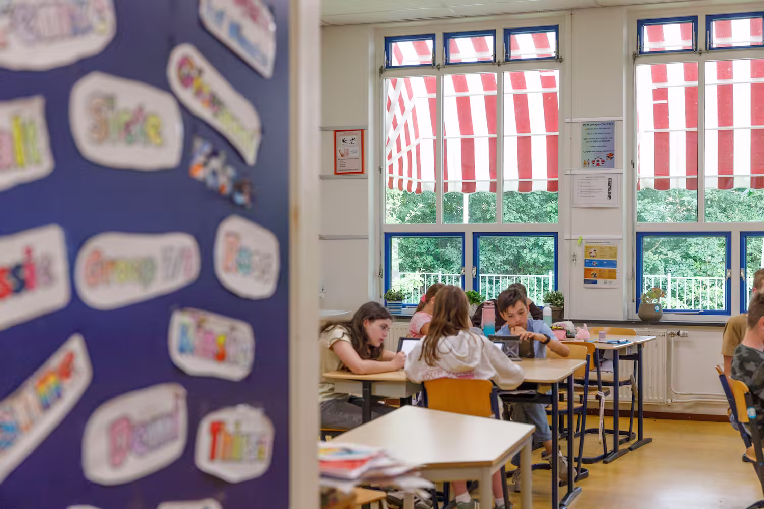 Kinderen zitten in een klaslokaal met grote ramen en rood-witte zonneschermen, werkend aan tafels.