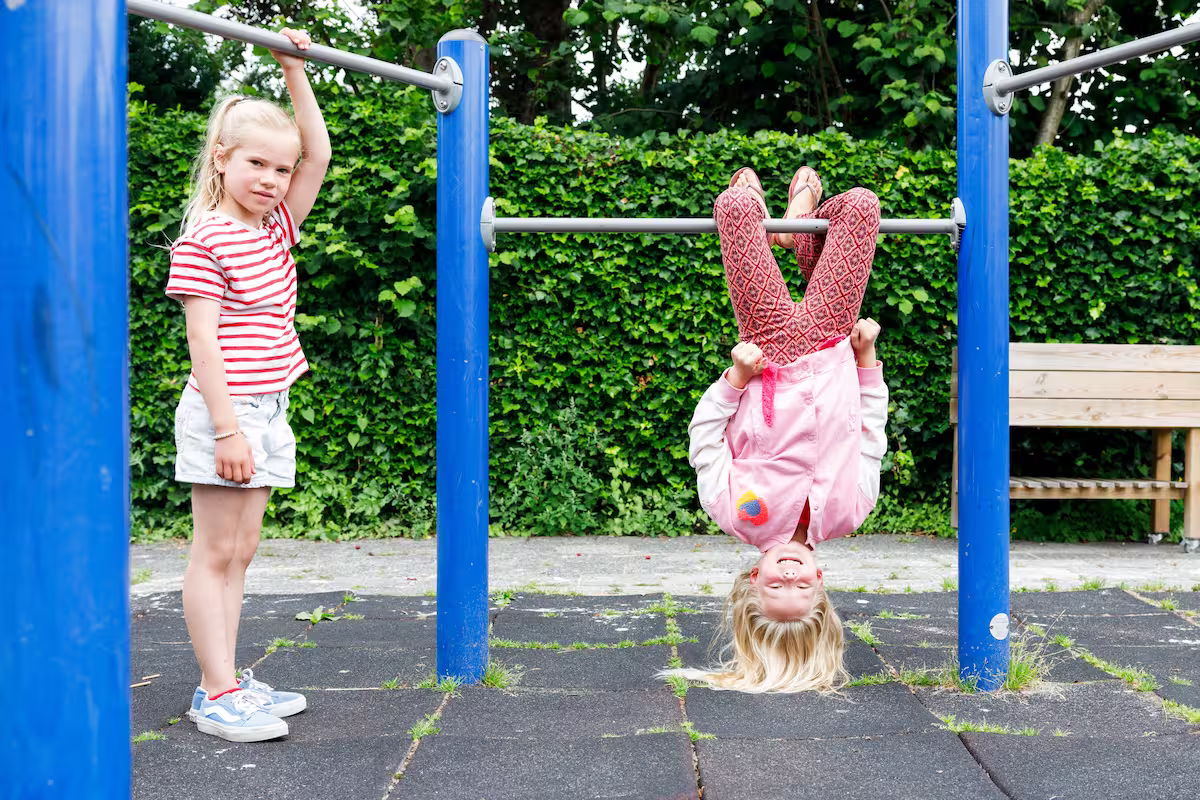 Twee meisjes spelen op een speeltoestel, de ene hangt ondersteboven aan een stang.
