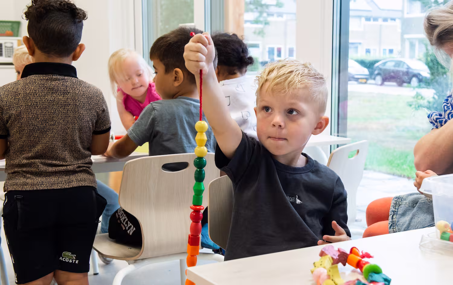 Jongen toont een rij gekleurde kralen aan een touw in een klaslokaal met andere kinderen.