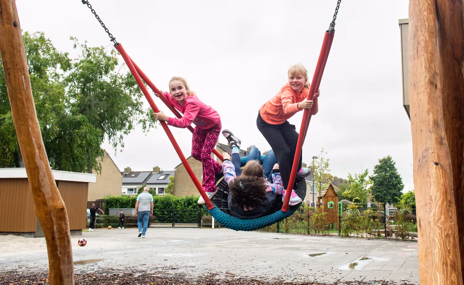 Vier kinderen zwaaien vrolijk op een grote schommel op een schoolplein met gebouwen op de achtergrond.