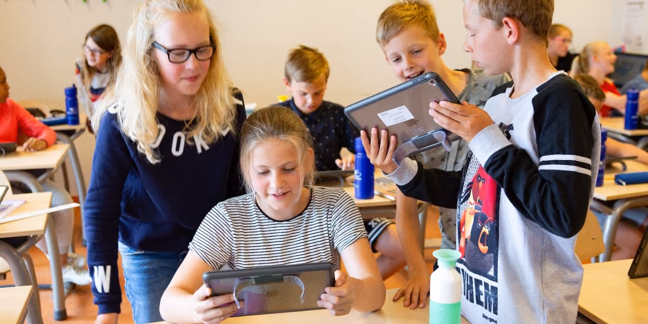 Vier kinderen bekijken tablets samen in een klaslokaal met andere leerlingen op achtergrond.