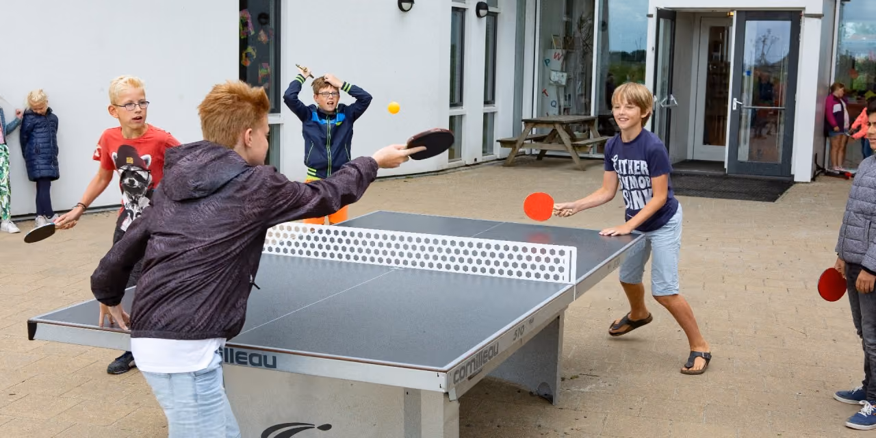 Kinderen spelen buiten tafeltennis op een tafeltennistafel, twee jongens slaan de bal met bats.