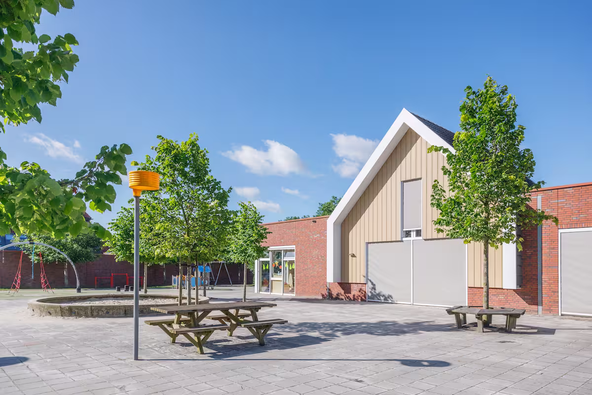 Buitenspeelplaats met speeltoestellen, picknickbanken en bomen naast een modern gebouw.