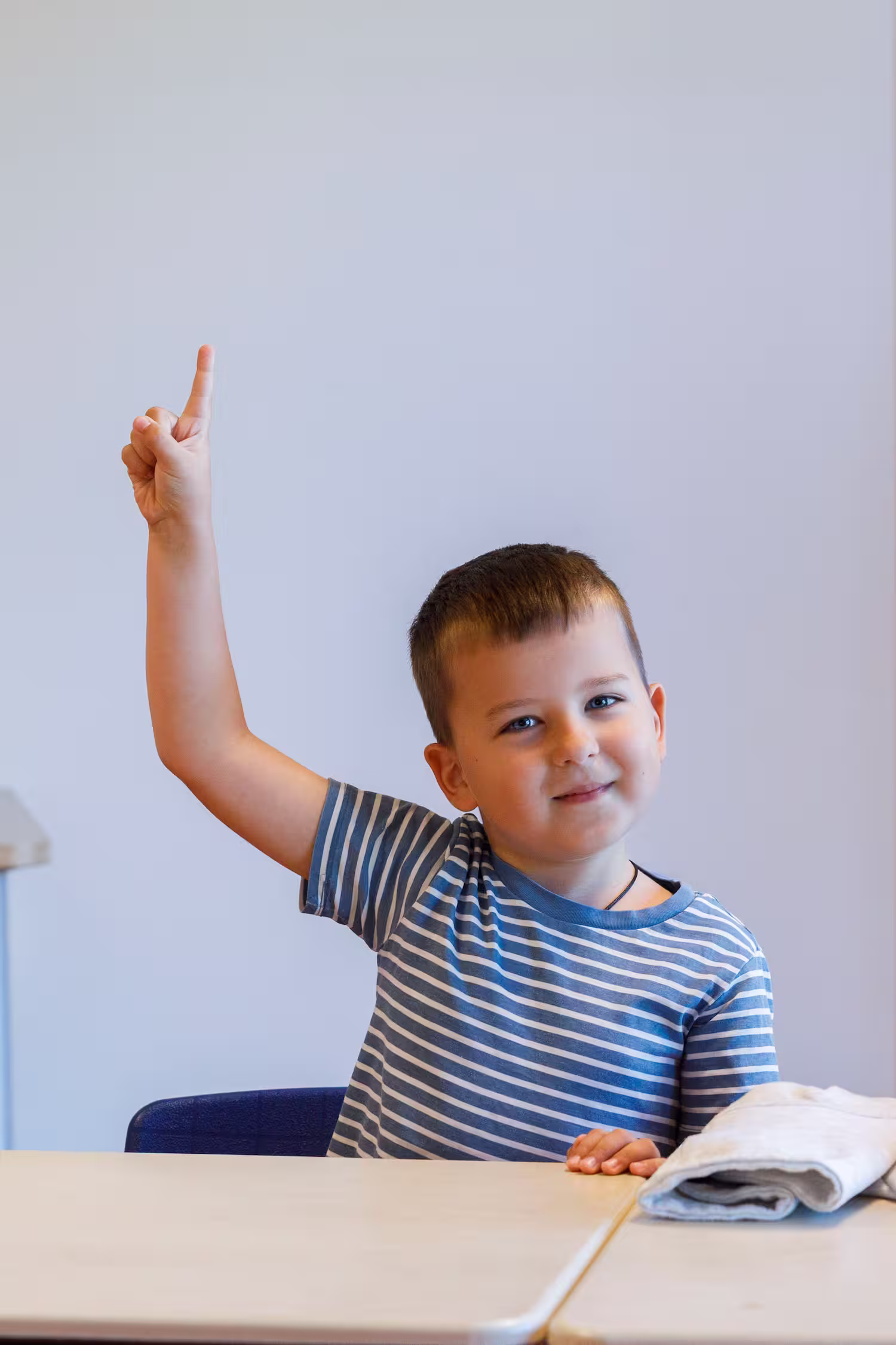 Jongen in gestreept shirt zit aan tafel en steekt vinger omhoog alsof hij een vraag heeft.