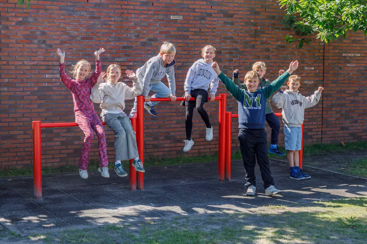 Zeven lachende kinderen spelen op en rond rood klimrek buiten tegen bakstenen muur.