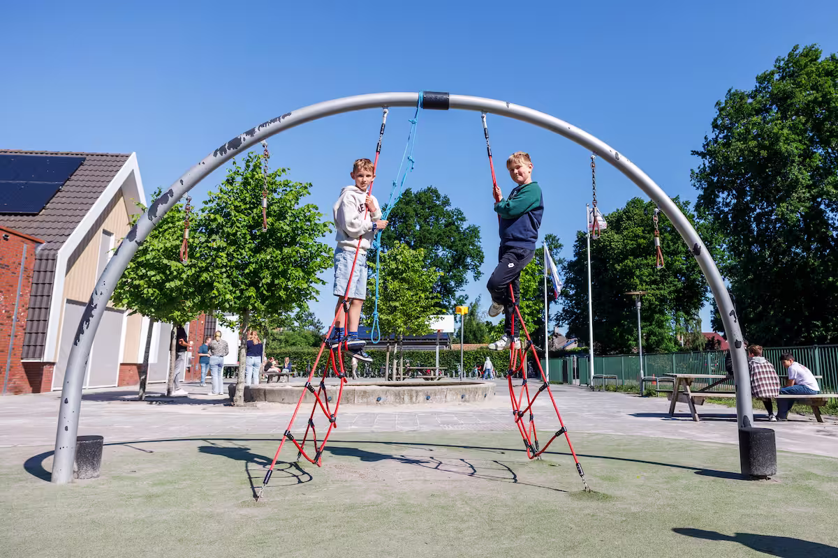Twee jongens klimmen aan rode touwen aan een metalen boog op een speelplaats onder een blauwe lucht.