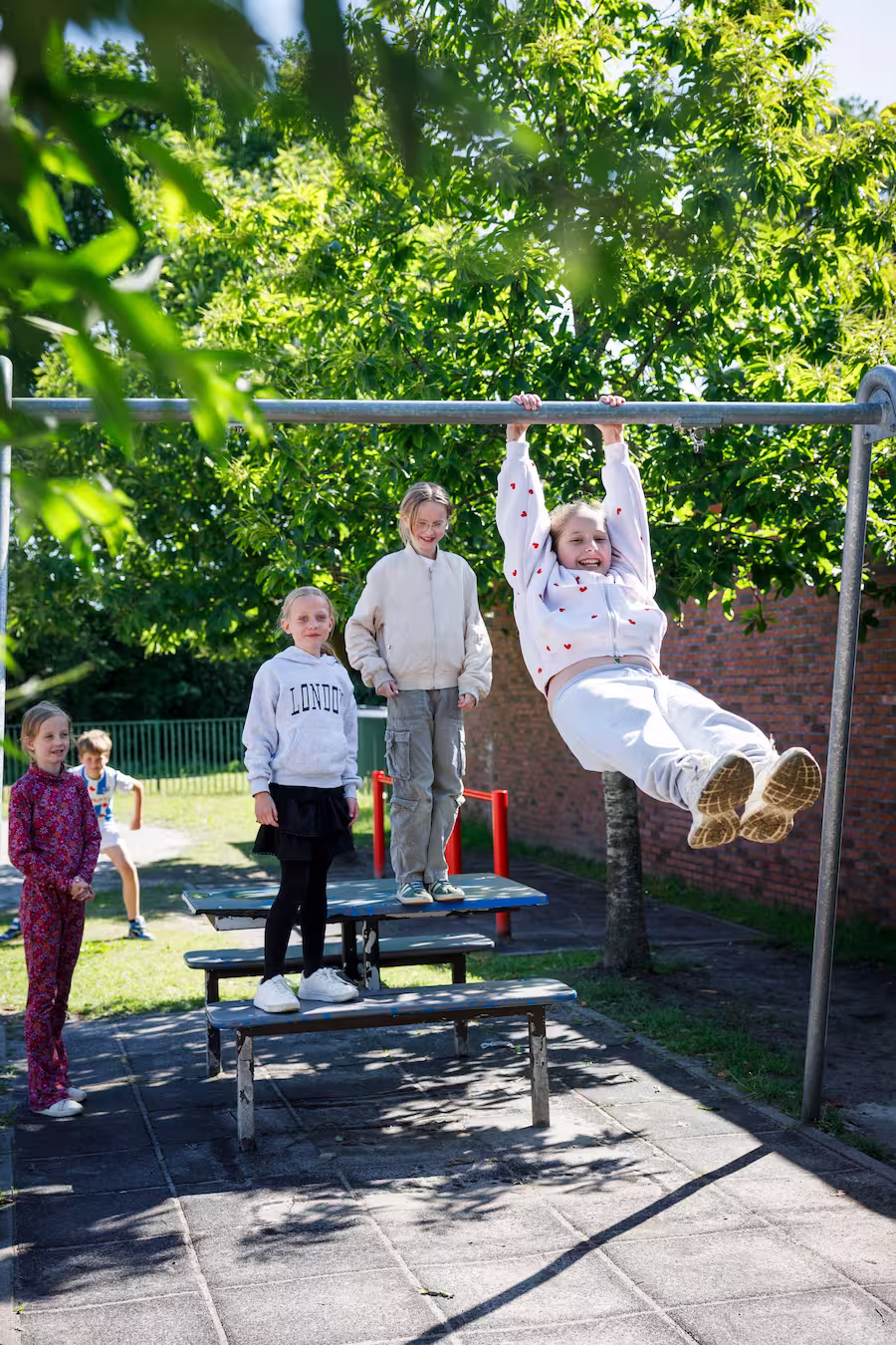 Kinderen spelen buiten; een meisje hangt aan een rek, anderen staan op een bankje in de zon.