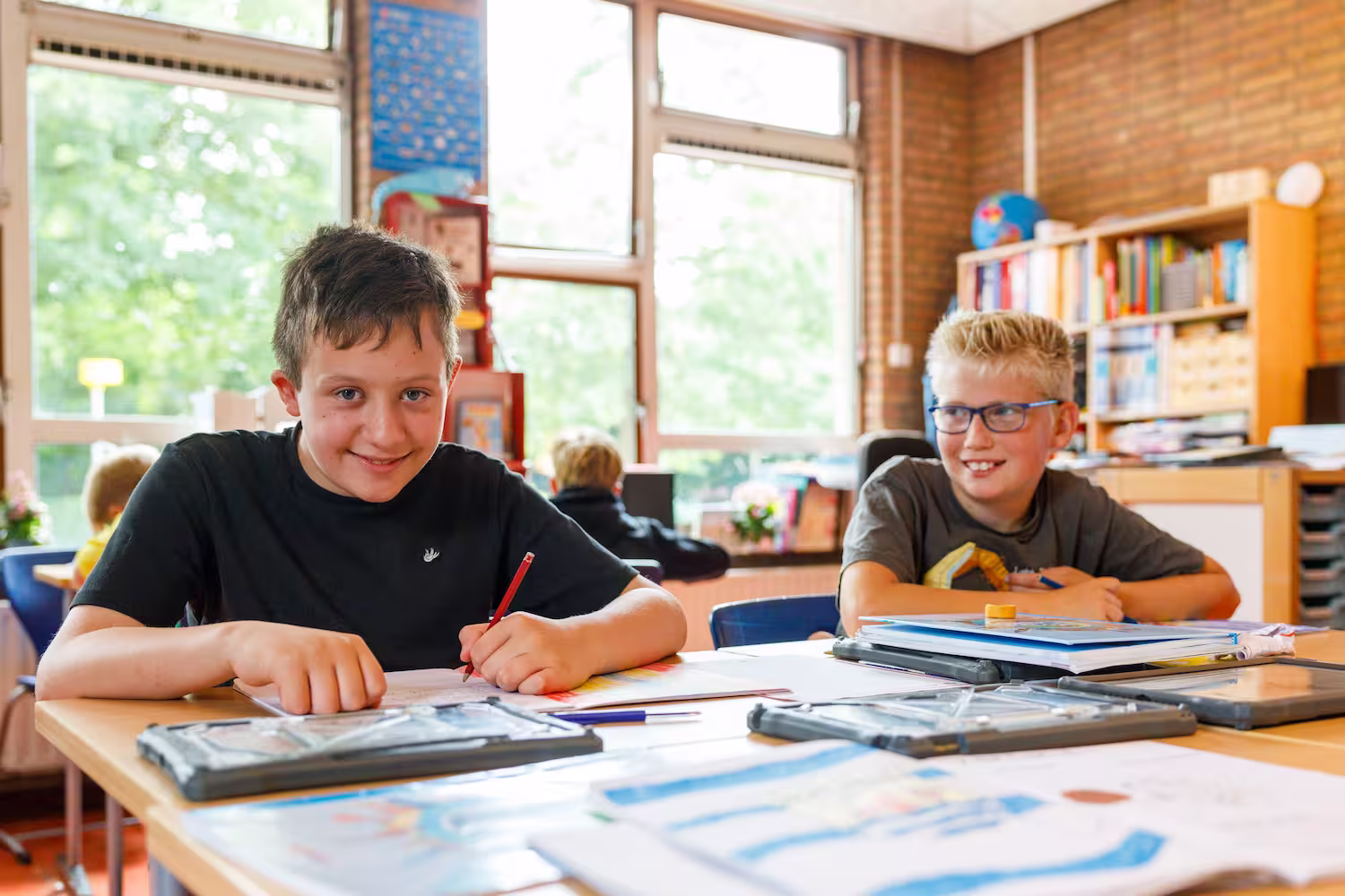 Twee jongens zitten aan een tafel in een klaslokaal, bezig met schoolwerk en tekenen.