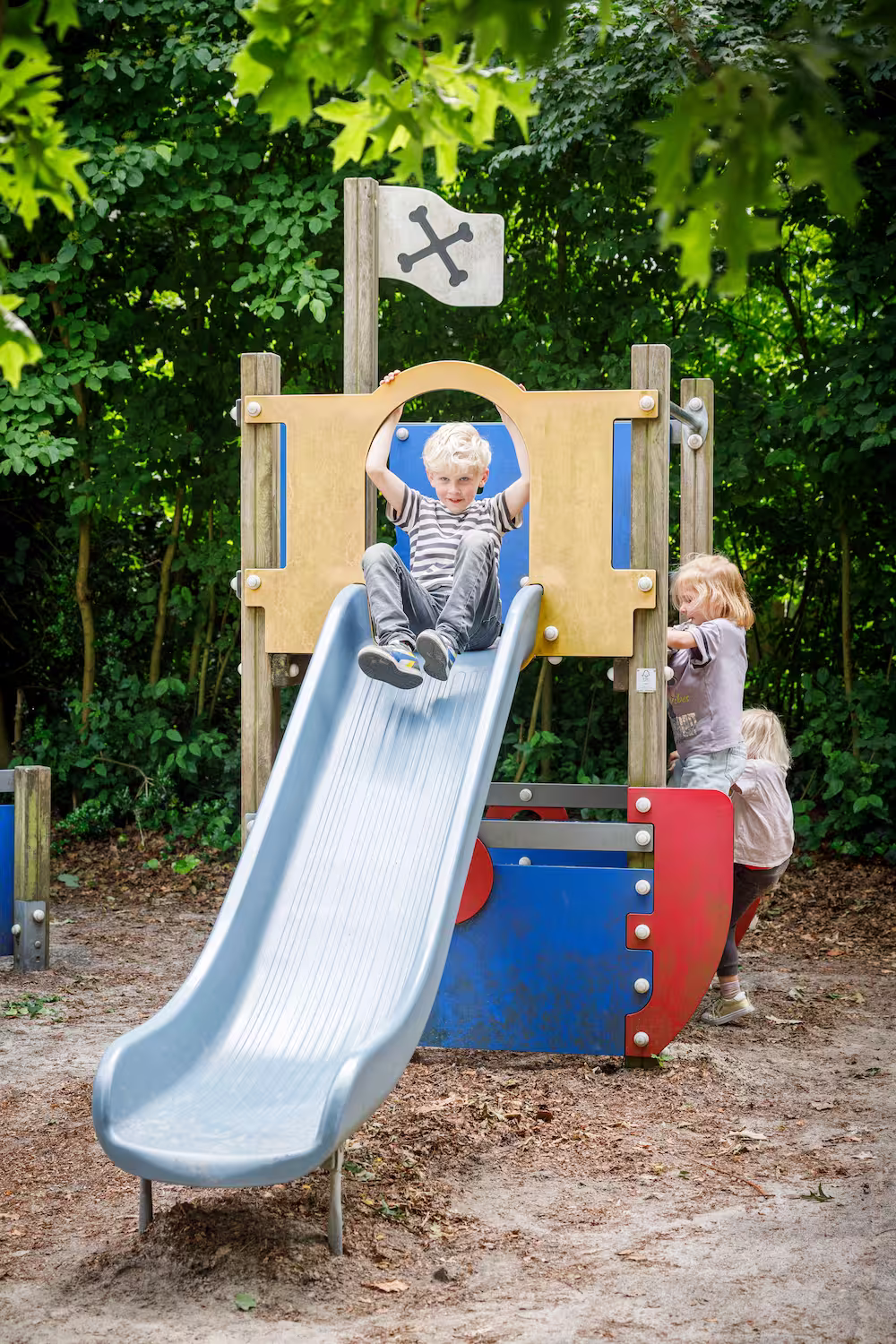 Blond jongetje glijdt van blauwe speeltoestelglijbaan terwijl twee kinderen klimmen, omgeven door groen.