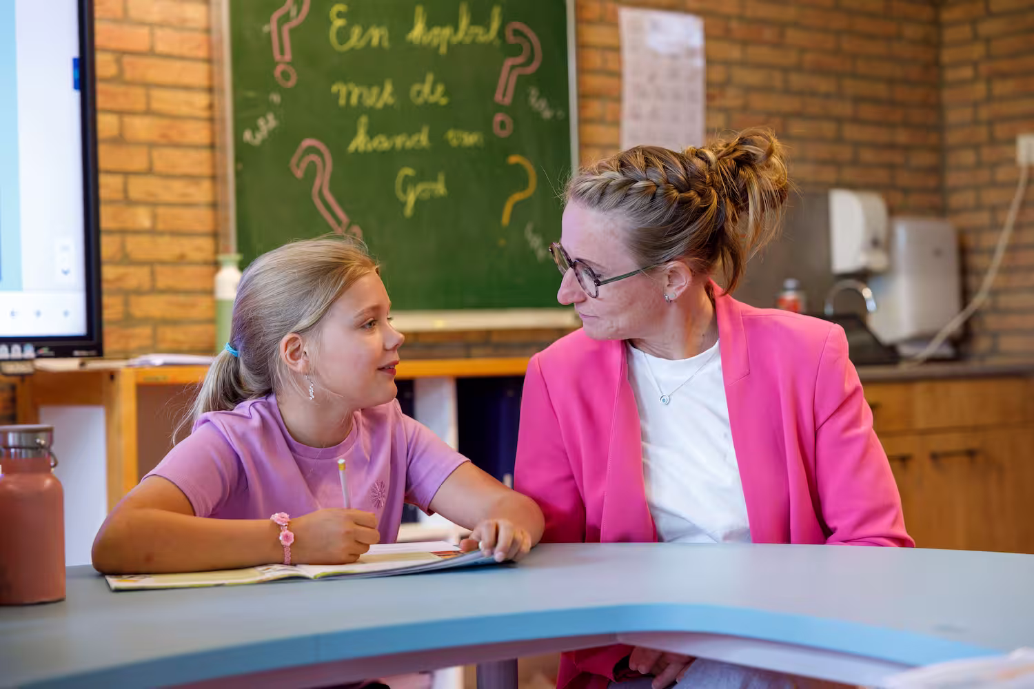 Meisje en vrouw praten aan een tafel in een klaslokaal met schoolbord op de achtergrond.