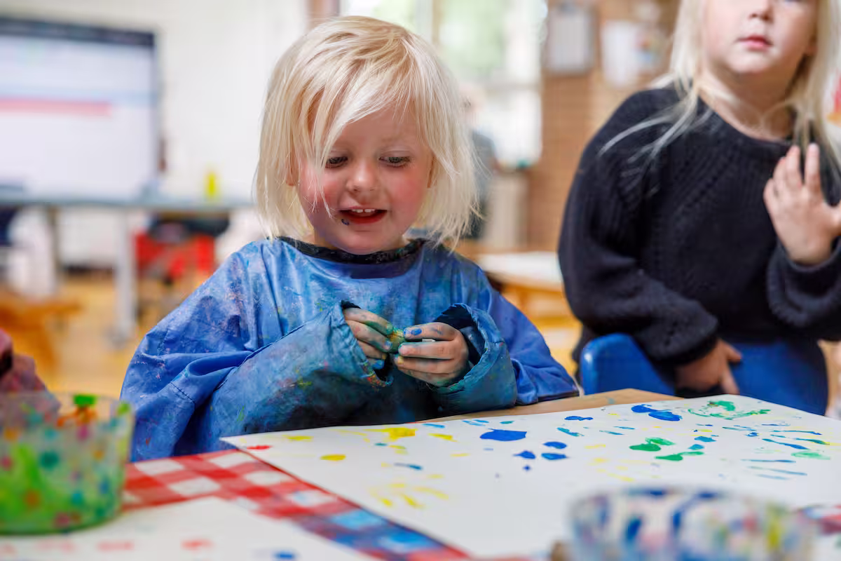 Klein blond kind draagt modderschort en schildert met felgekleurde verf op papier.