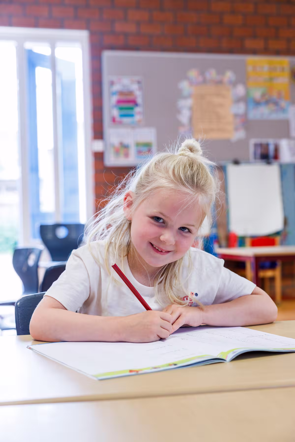 Blond meisje zittend aan een tafel, schrijf met rood potlood in een open schrift in een klaslokaal.