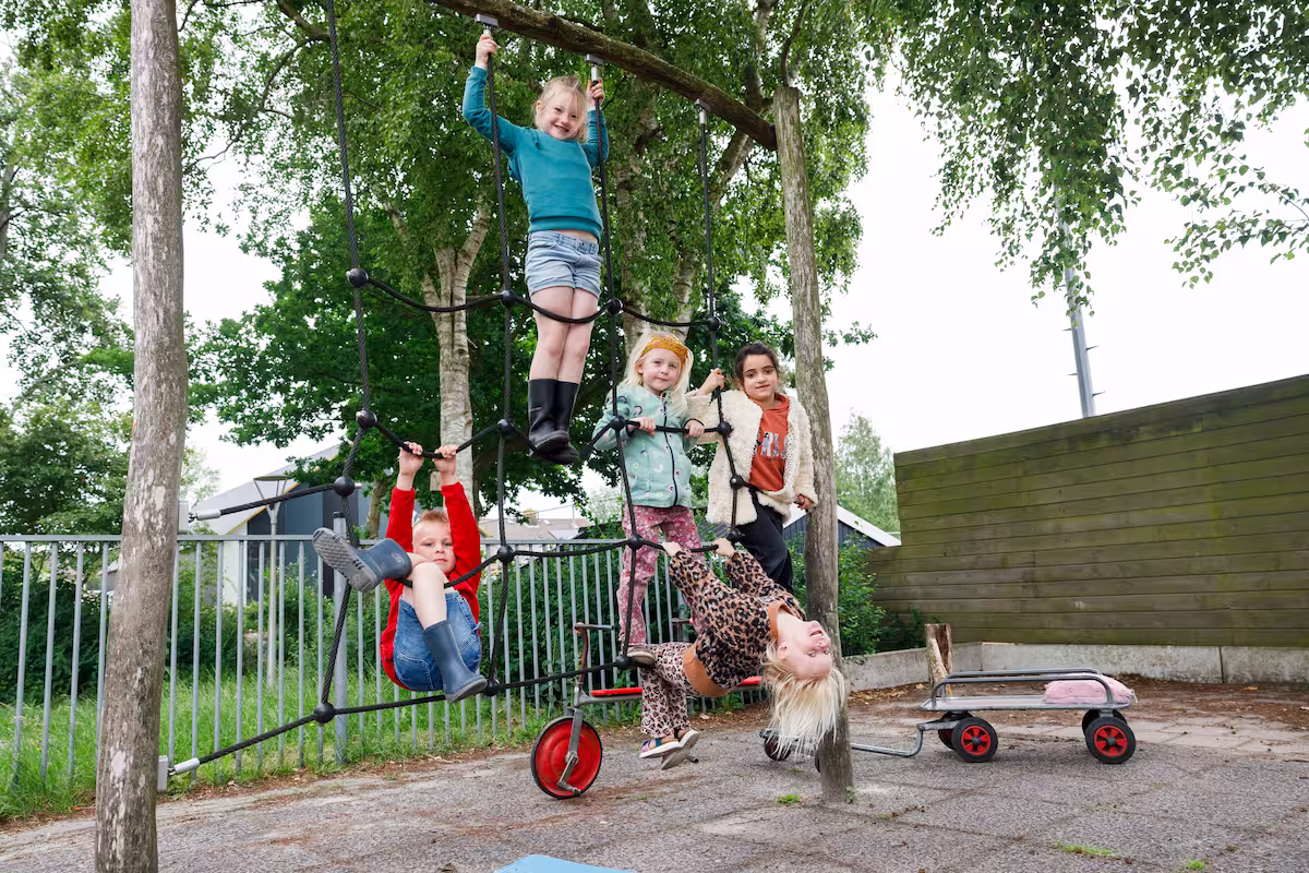 Vijf kinderen spelen lachend samen in een klimnet op een buitenspeelplaats.