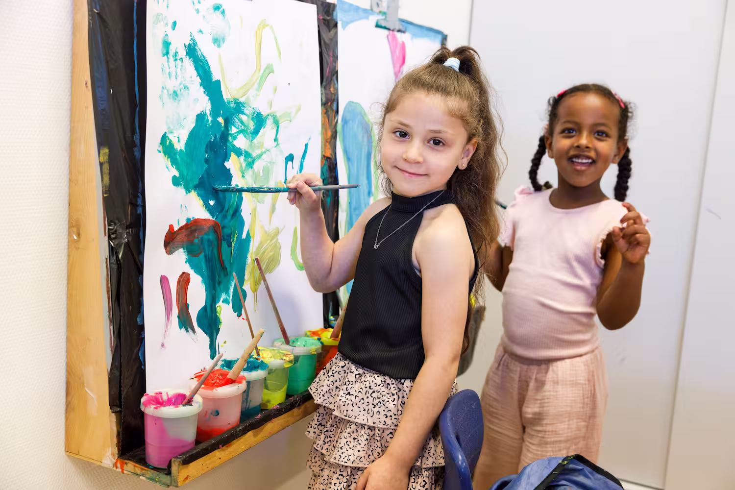 Twee meisjes schilderen aan een muur met kleurrijke verf en kijken naar de camera.