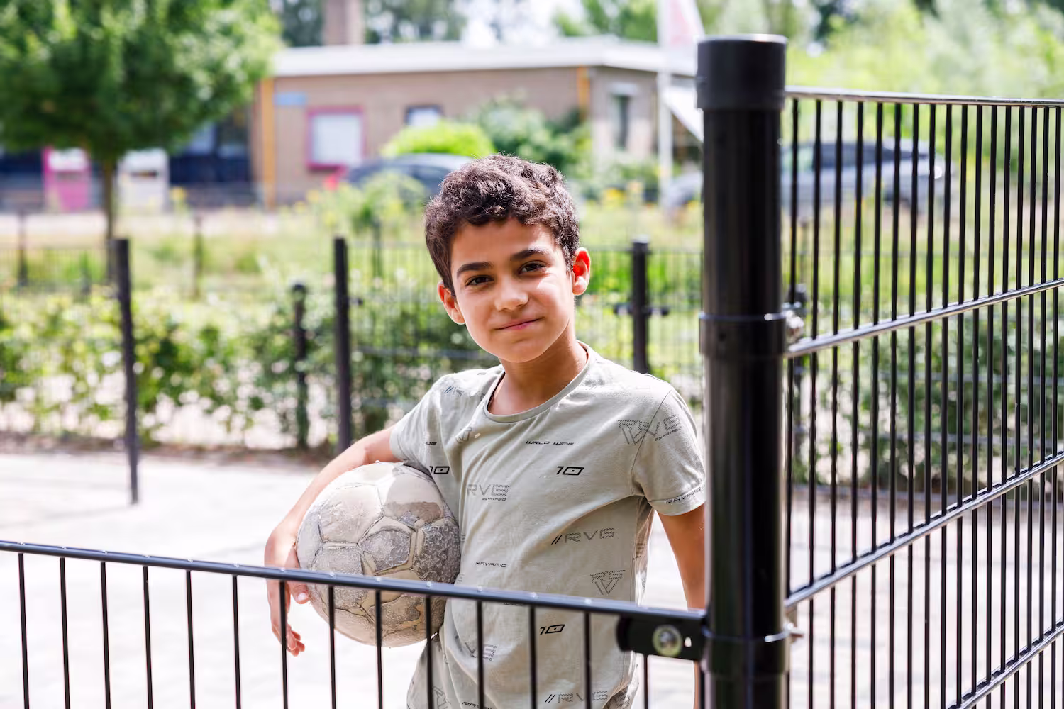 Jongen met een bal leunt ontspannen op een hek bij een speelplaats buiten.
