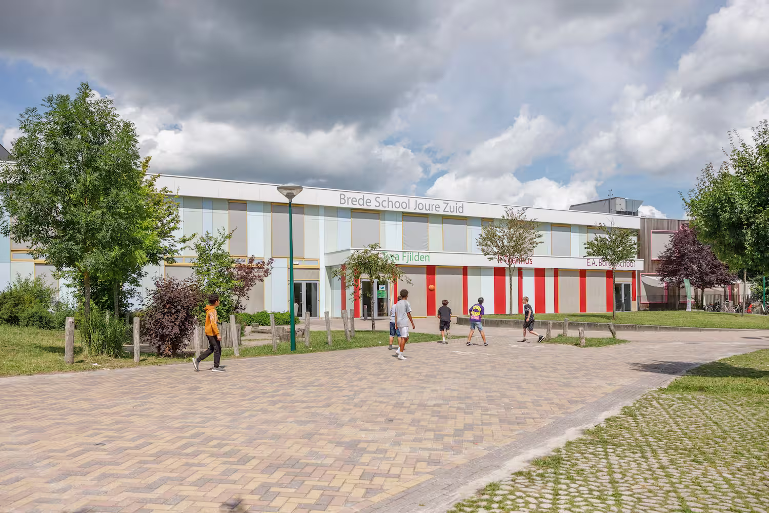 Kinderen spelen buiten voor het gebouw van Brede School Joure Zuid op een zonnige dag met wolkenlucht.