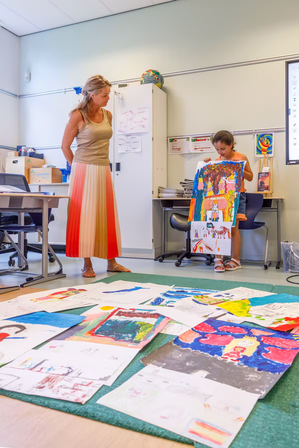 Meisje toont kleurrijke tekeningen aan een vrouw in een klaslokaal met kunstwerken op de vloer.