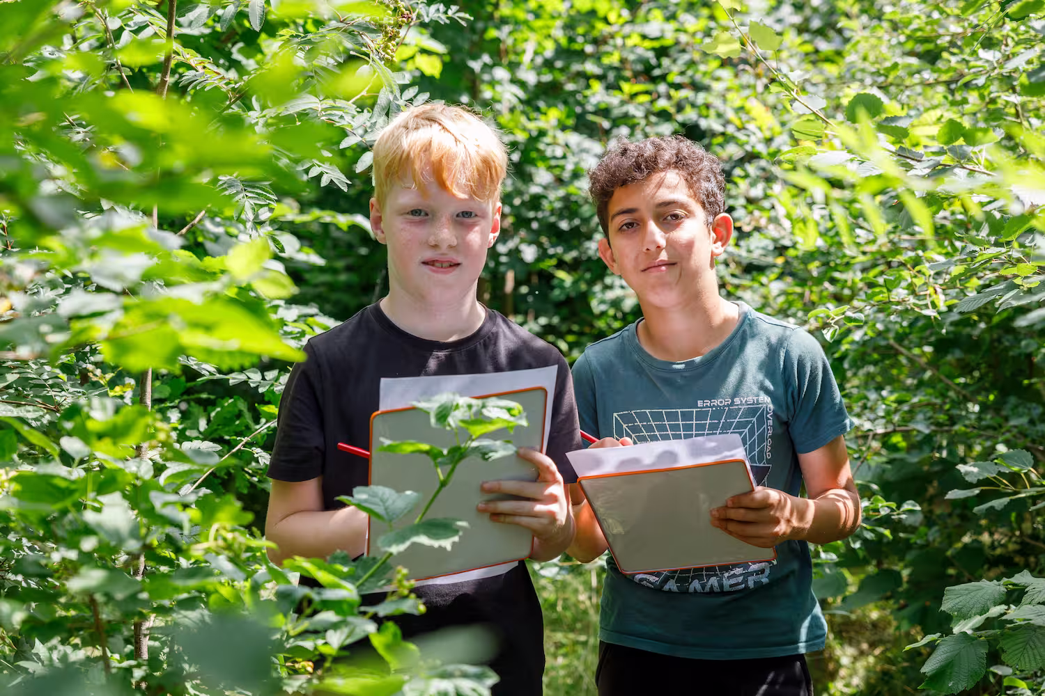 Twee jongens staan in groene natuur met clipboards en potloden, bezig met aantekeningen maken.