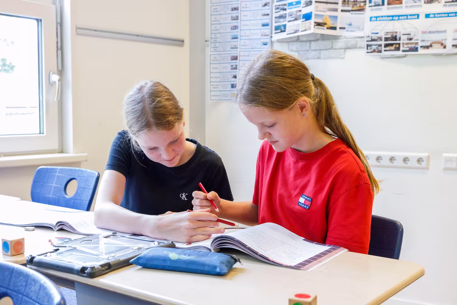 Twee meisjes zitten in een klaslokaal en werken samen in een open boek met rode potloden.