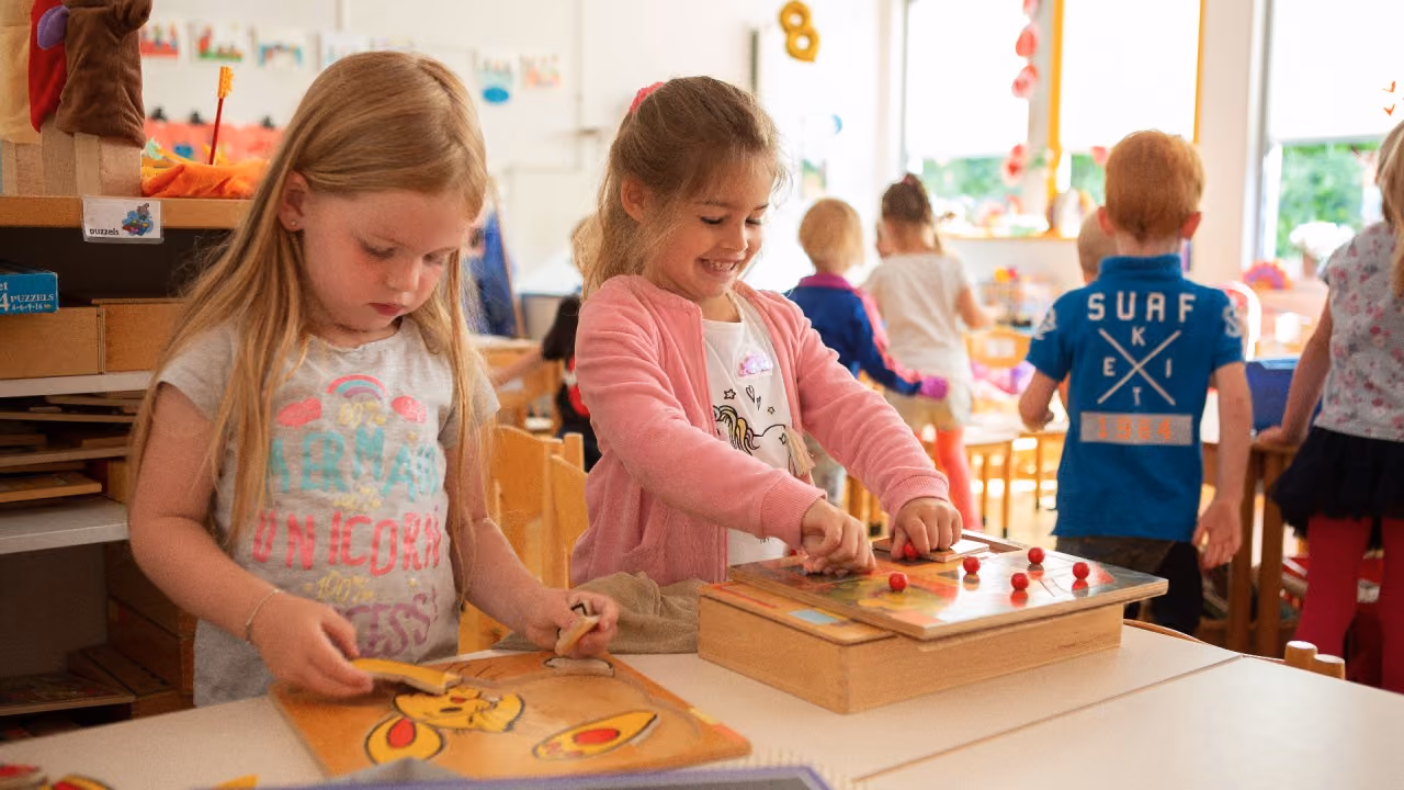 Twee meisjes spelen met houten puzzels aan een tafel in een klaslokaal.