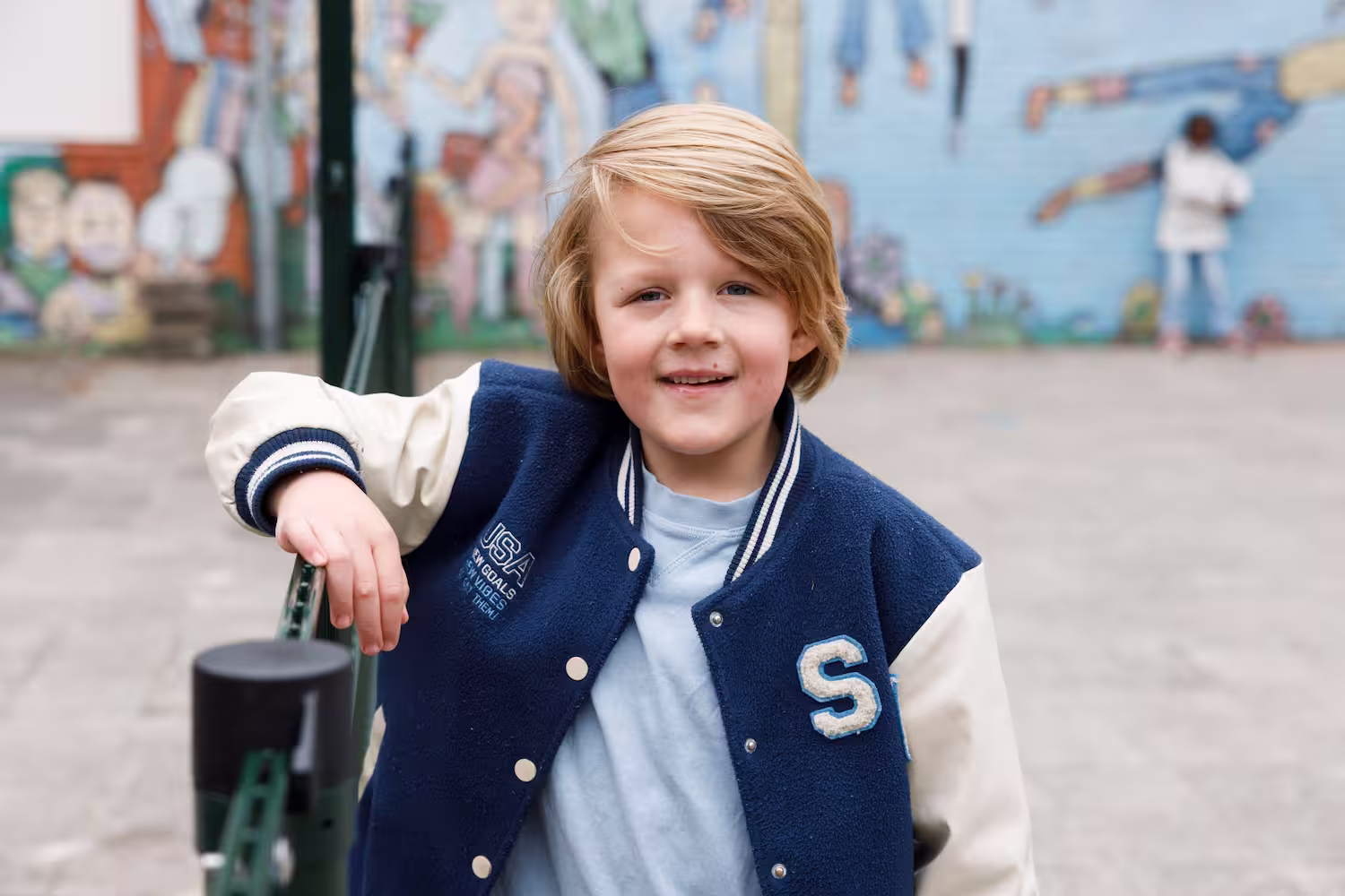 Jongen met blond haar en blauw-witte varsity jas leunt op groen hek buiten met muurschildering op achtergrond.