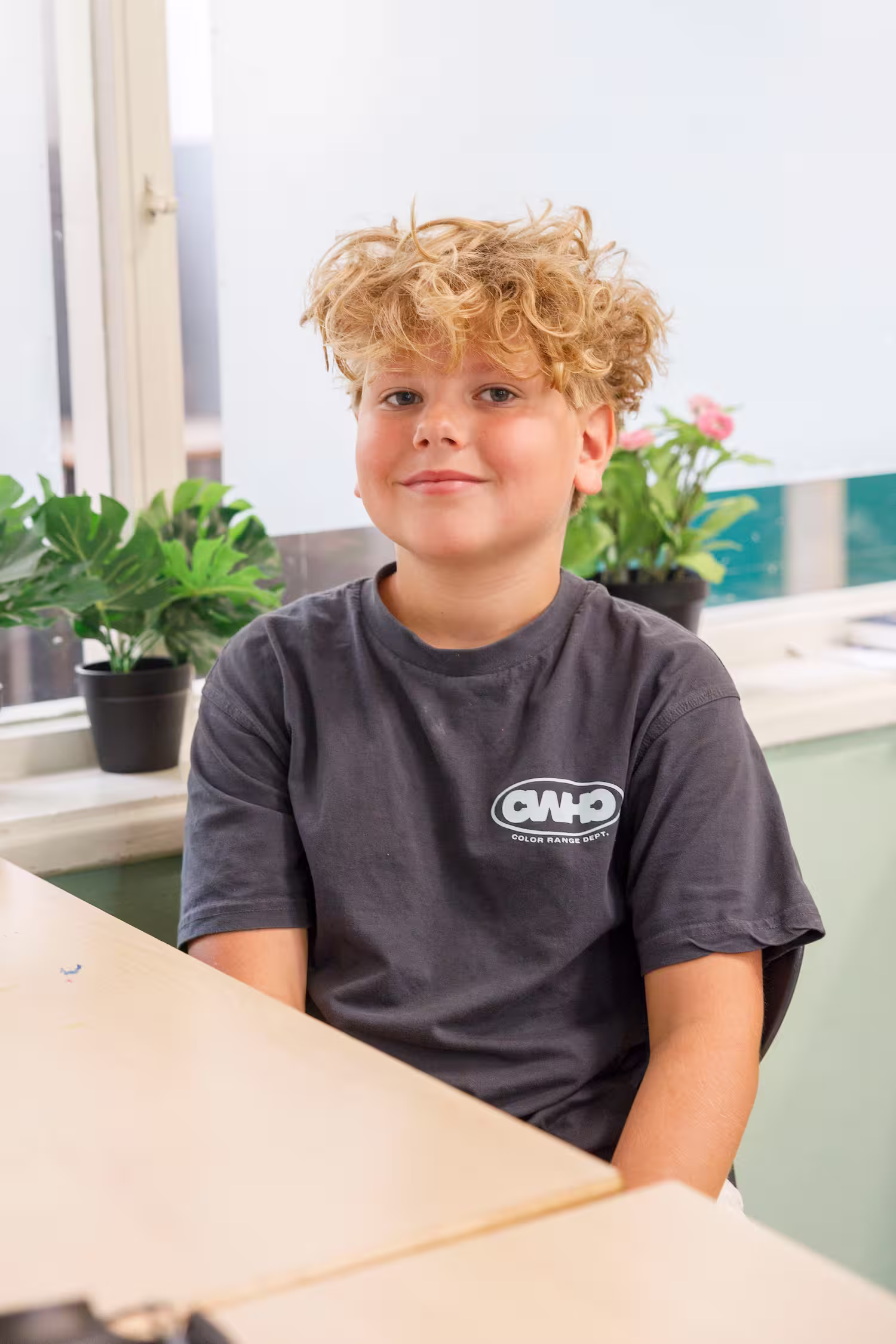 Jongen met krullend blond haar die in een kamer aan een tafel zit, met planten op de vensterbank achter hem.
