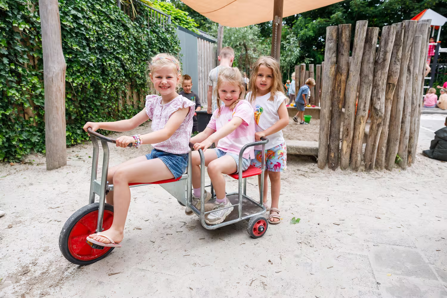 Drie meisjes spelen op een driewieler in een zandbak op een speelplaats met houten palen.