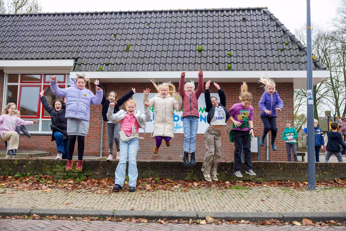 Kinderen springen vrolijk van een lage muur voor een gebouw met een zwart dak en rode ramen.