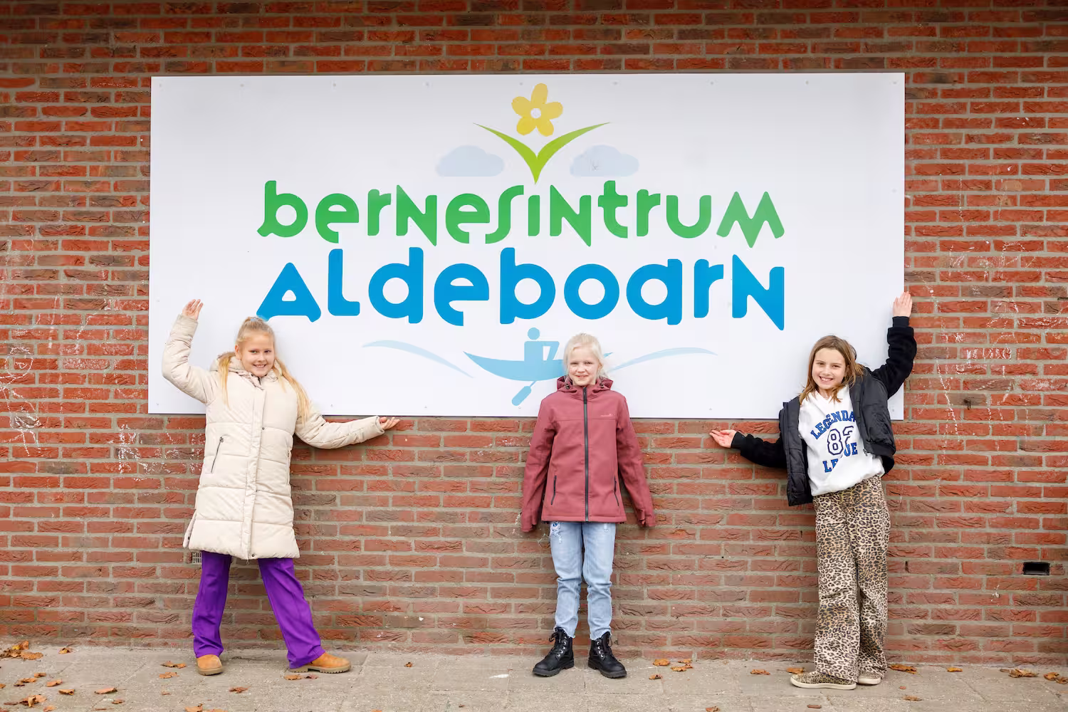 Drie kinderen poseren voor een bakstenen muur met een bord 'Bernsintrum Aldeboarn'.